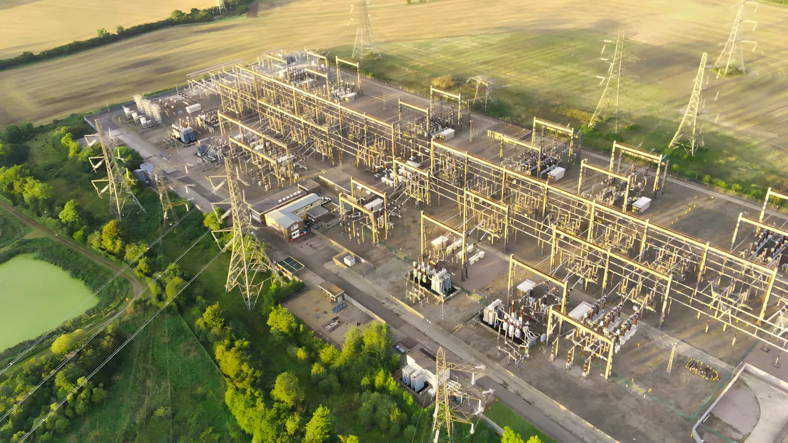Aerial view of a high-voltage substation, with rows of transformers, busbars and gantries, and overhead line towers running out across farmland.